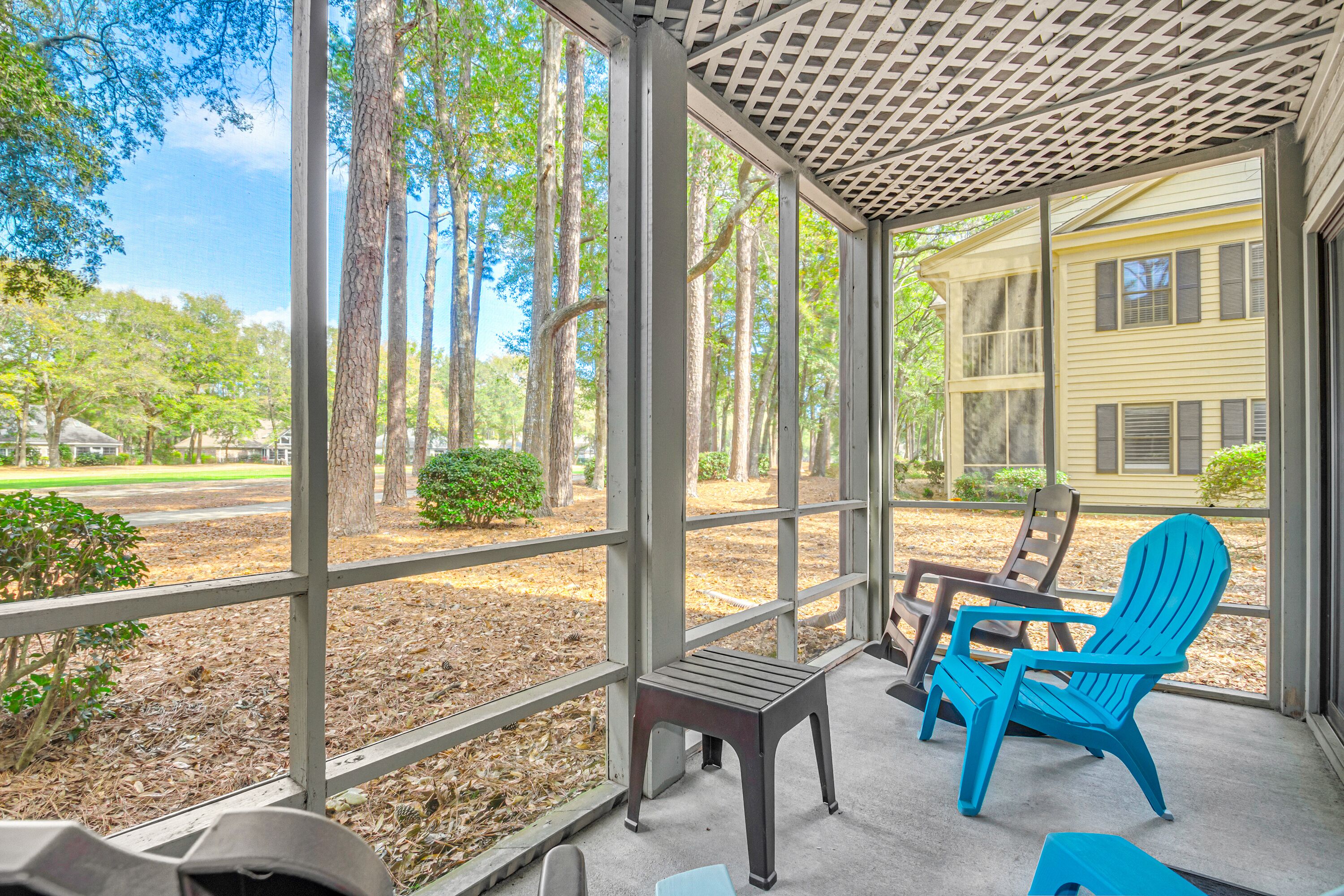 A covered porch with a view of trees and a building. There are two Adirondack chairs and a small table.
