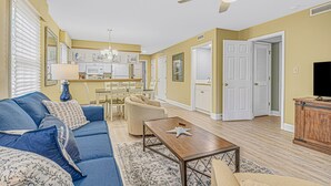 A living room with a blue sofa, beige armchair, and a wooden coffee table. There is a ceiling fan, a kitchen in the background, and a television on a wooden cabinet. - PP 200-2 Golfers Paradise in Pawleys Plantation (Pawleys Island)