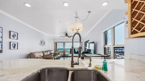 A modern kitchen with a double sink, a hanging light fixture, and a view of the living area with a sofa and a pool. - Emerald Towers 1007 * Beach Chair Set Included * Salt Water Vacations (Fort Walton Beach)