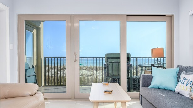 A modern living room with a sofa, a table, and a balcony with a view of the beach. - Emerald Towers 1007 * Beach Chair Set Included * Salt Water Vacations (Fort Walton Beach)