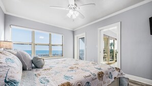 A bedroom with a large bed, a ceiling fan, a view of the sea through a window, and a door leading to a balcony. - Emerald Towers 1007 * Beach Chair Set Included * Salt Water Vacations (Fort Walton Beach)