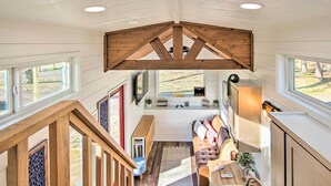 Un loft espacioso con techos altos, vigas de madera, una zona de estar acogedora con sofá y banco, y grandes ventanales con vistas al exterior.