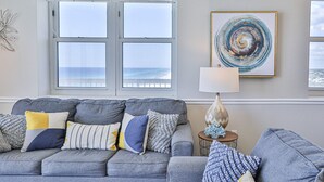 A grey sofa with multiple cushions, a small table with a lamp, and a framed artwork on the wall. - Gulf front end unit w/ wraparound balcony, Sunset ocean views, 3BR 3BA! (Panama City Beach)