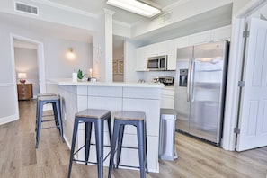 A modern kitchen with white cabinets, stainless steel appliances, and a central island with stools. - Gulf front end unit w/ wraparound balcony, Sunset ocean views, 3BR 3BA! (Panama City Beach)
