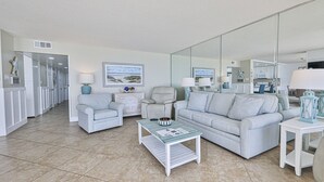 En rummelig stue med en lys sofa, lænestole, et sofabord og et stort spejl der reflekterer rummet.