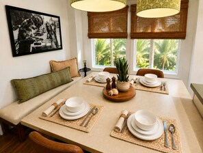 Zona pranzo con tavolo apparecchiato per quattro persone, divano, quadro appeso al muro e vista su palme attraverso la finestra.