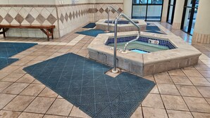 A pool area with two hot tubs, blue mats, a bench, and a tiled floor.