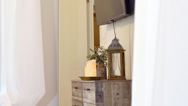 A bedroom with a wooden dresser, a mirror, a television mounted on the wall, and a bedside table with a lamp. - Relaxing Cottage Near Fishhawk (Lithia)