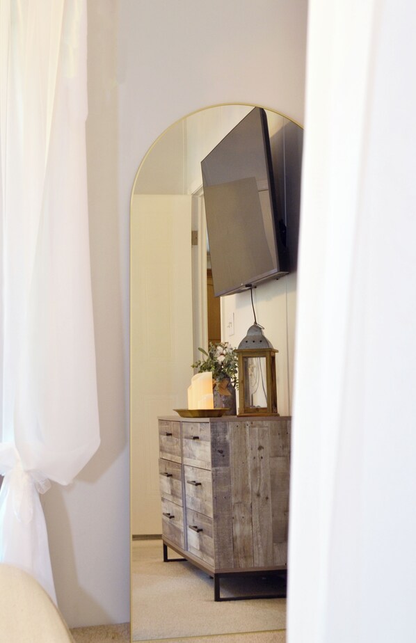 Une chambre à coucher avec une commode en bois, un miroir, un téléviseur fixé au mur et une table de chevet avec une lampe.