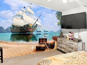 A bedroom with a large mural of a sailing ship on a beach, a wooden dresser, and a television.