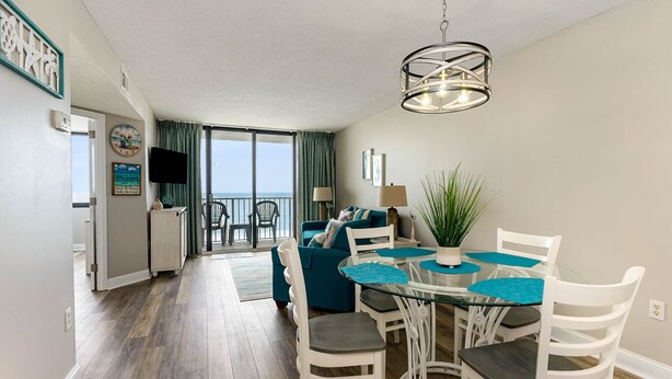 Een eethoek met een glazen tafel en witte stoelen, een blauwe bank en uitzicht op de oceaan door grote ramen.