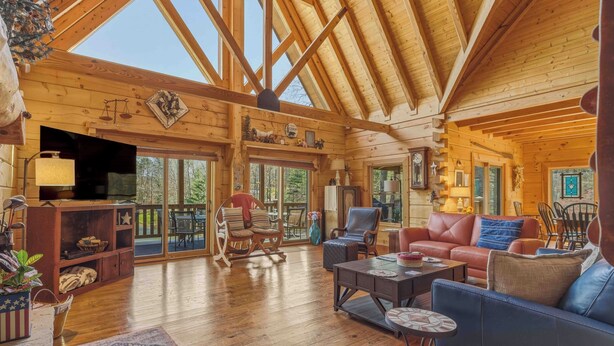 Un intérieur spacieux de chalet en bois, avec un plafond haut, des poutres apparentes, une cheminée et une grande porte-fenêtre coulissante donnant sur un espace extérieur.