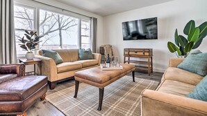 Ein Wohnzimmer mit einer Couch, einem Hocker und einem Couchtisch. Ein Fernseher ist an der Wand montiert und es gibt ein großes Fenster mit Vorhängen.