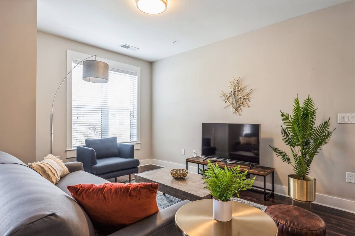 A modern living room with a grey sofa, a blue armchair, a round coffee table, a flat-screen TV, and a large window with blinds.