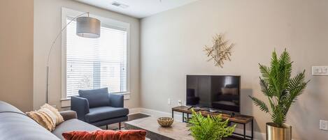 A modern living room with a grey sofa, a blue armchair, a round coffee table, a flat-screen TV, and a large window with blinds.