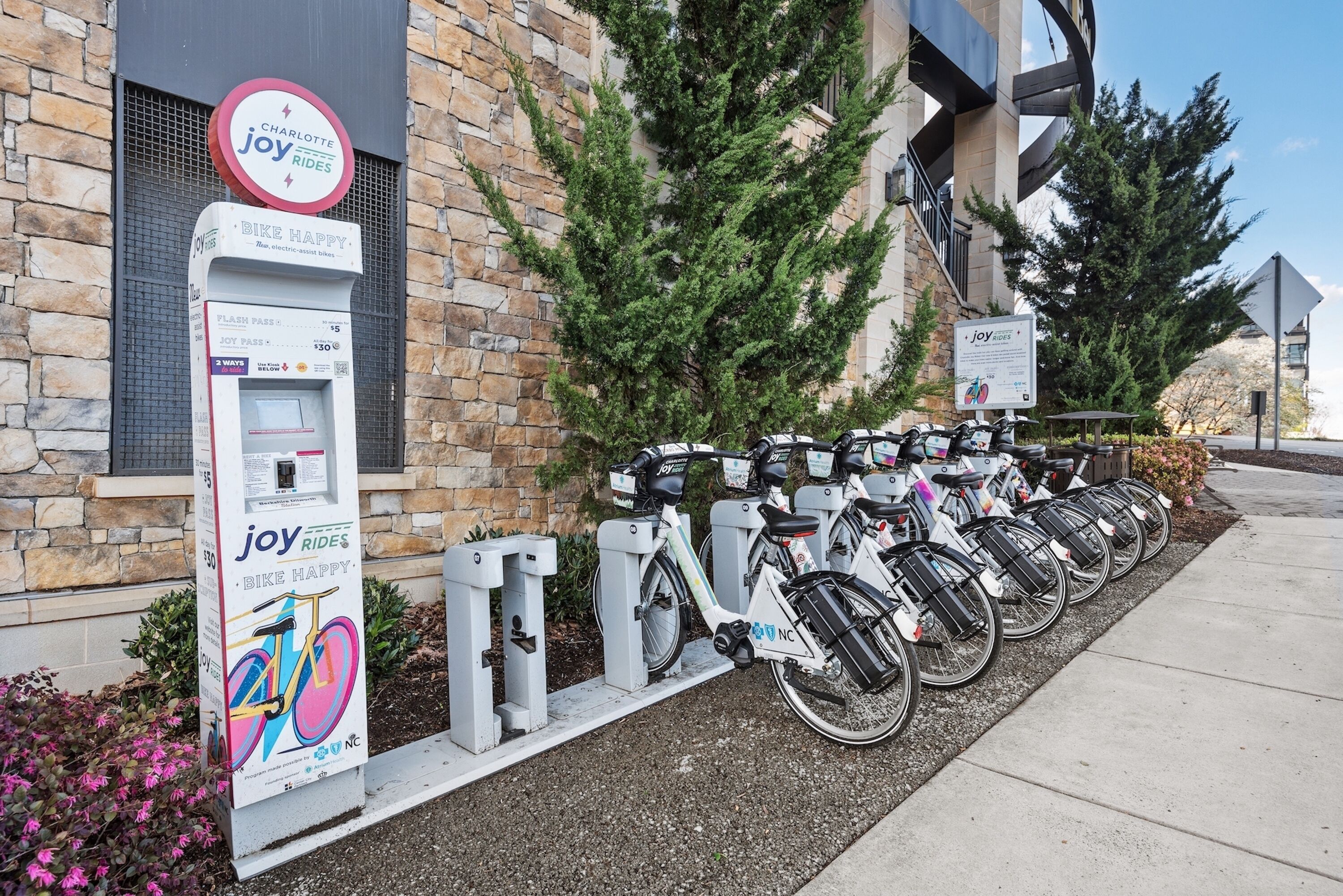 A bike rental station with a 'Joy Rides' sign, a bike charging station, and a row of parked bicycles.