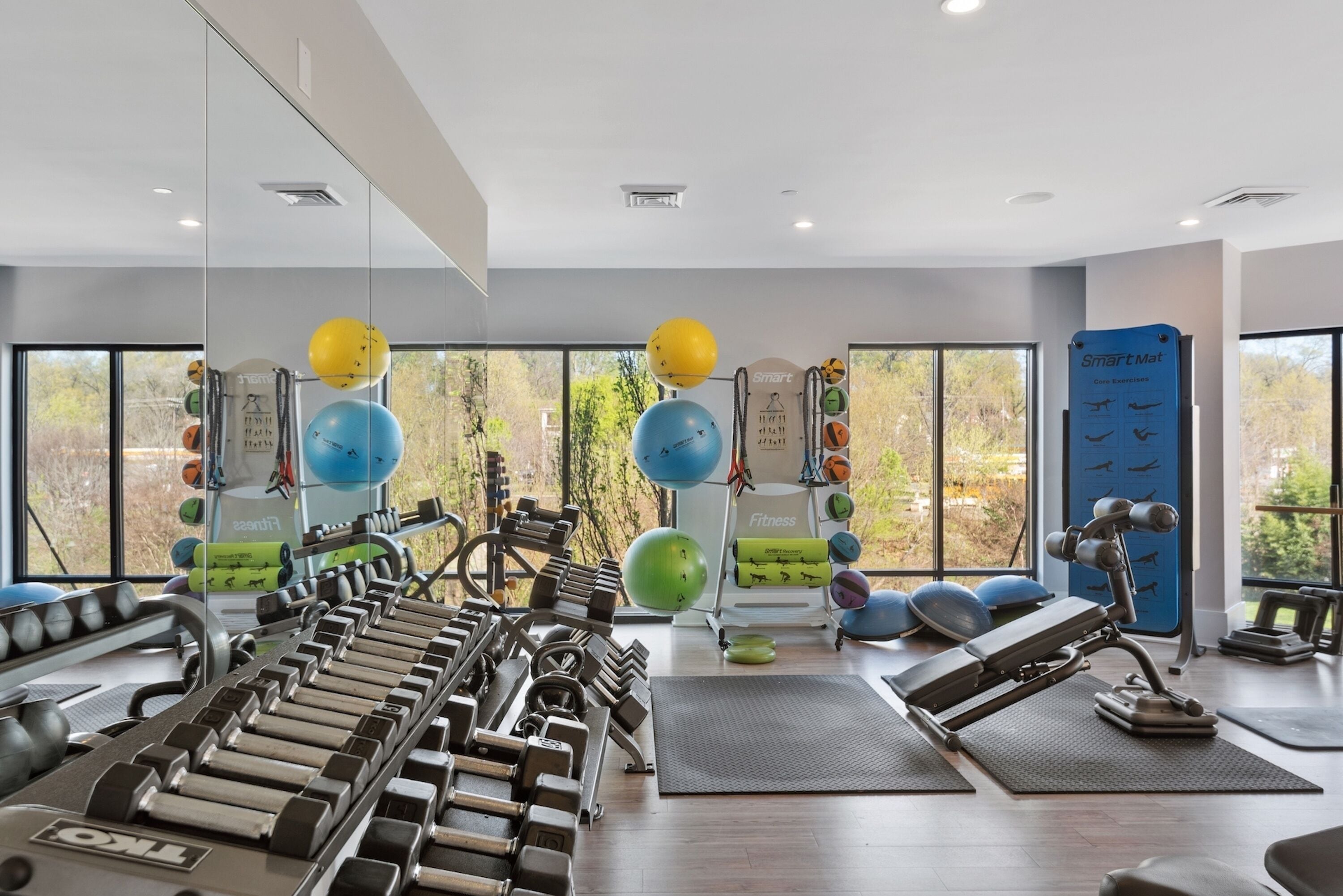A modern gym with various exercise equipment, large windows, and a view of trees.