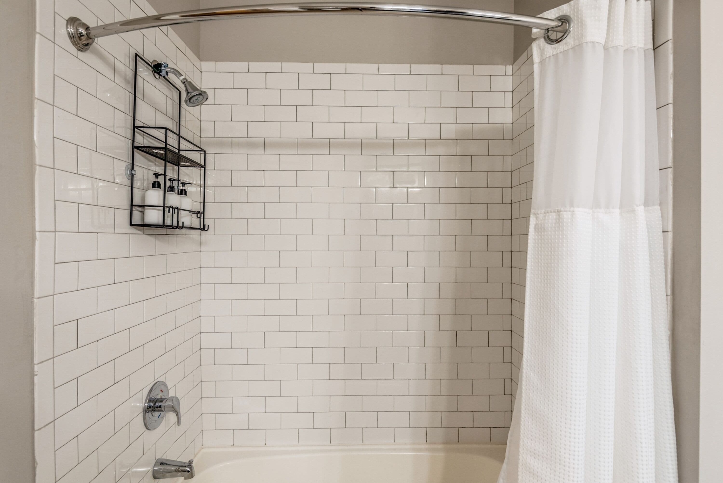A shower area with white tiled walls, a chrome showerhead, a glass shower caddy, and a white shower curtain.
