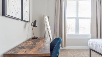 A room with a wooden desk, a blue chair, a bed, and a window with curtains.