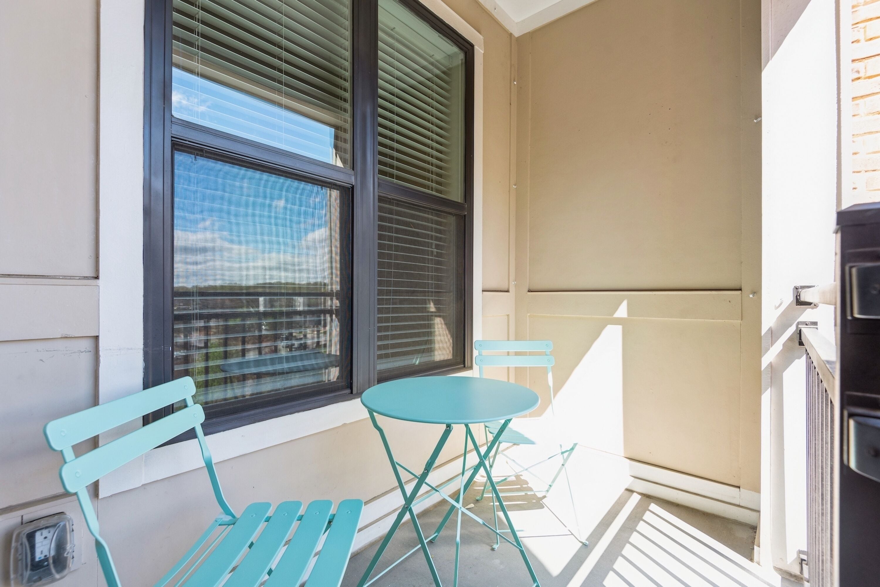 A balcony with a table and two chairs, a window with blinds, and a door to the right.