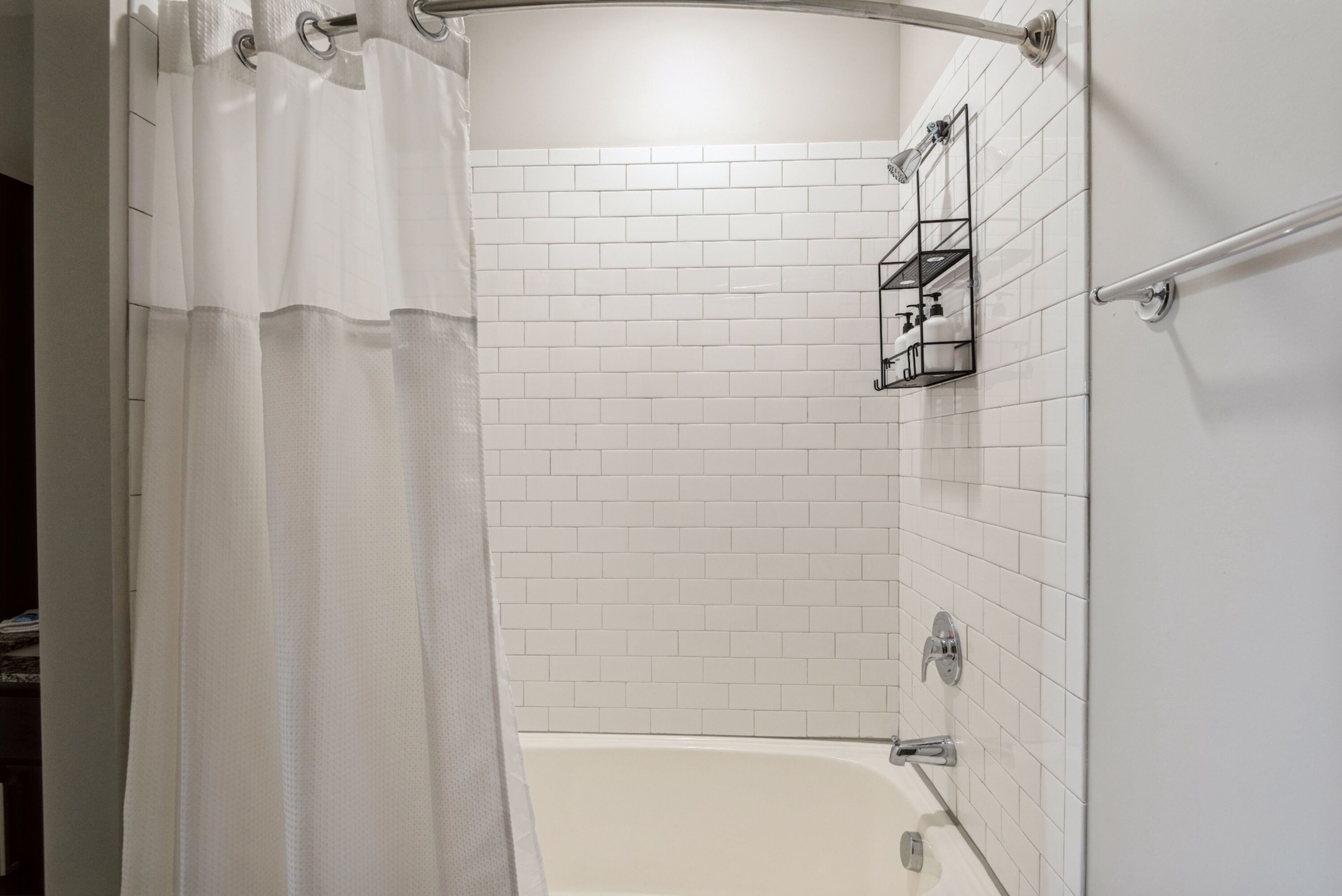 A bathroom with white tiled walls, a bathtub, a shower with a curtain, and a shelf with toiletries.