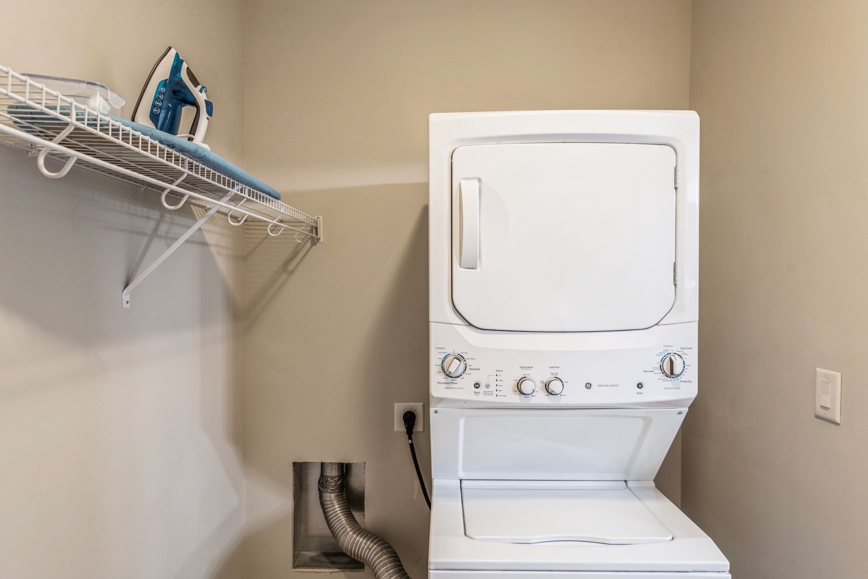 A white washing machine with a control panel, a metal drying rack, and a blue spray bottle on a shelf.