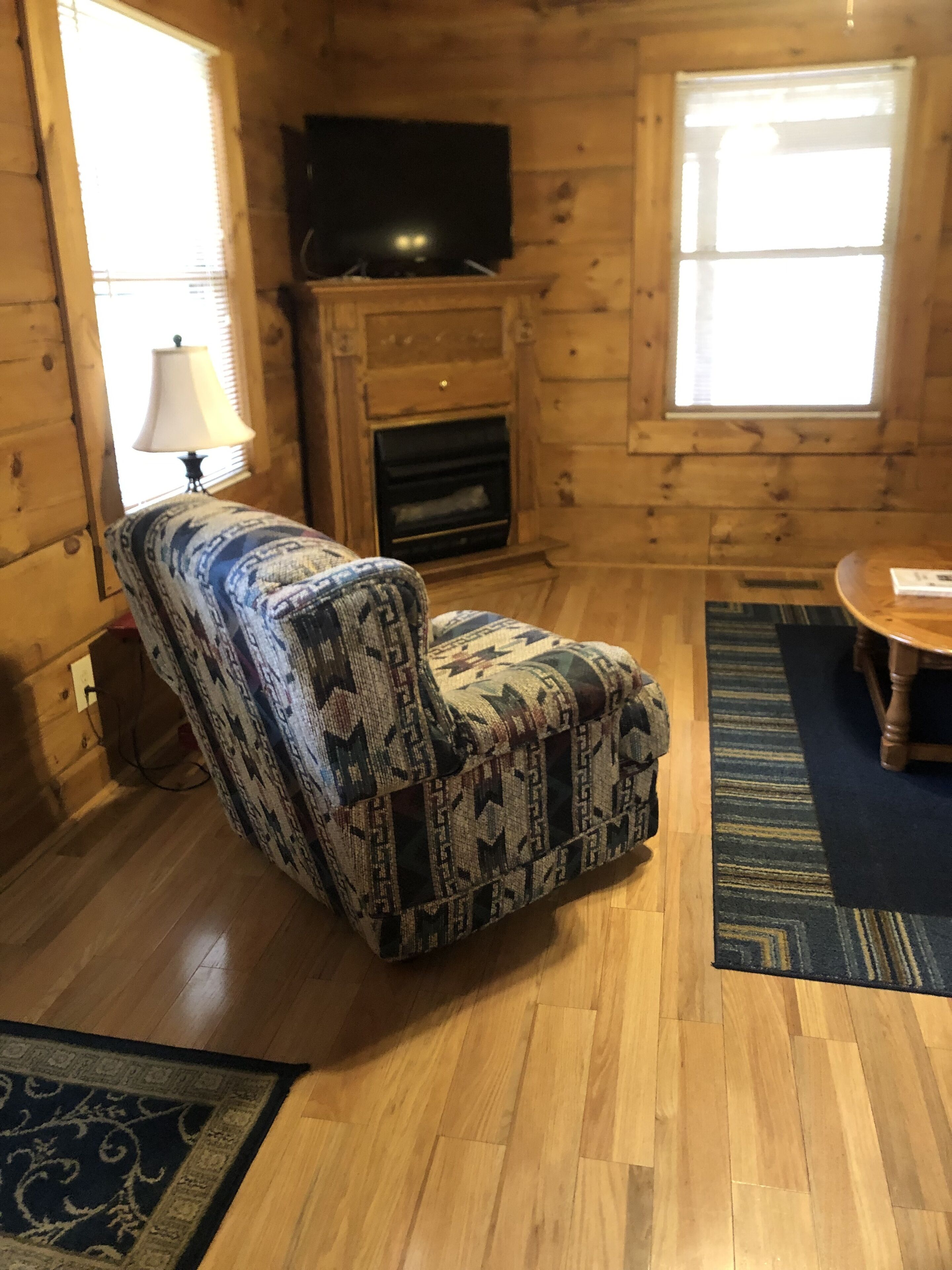A living room with a patterned armchair, a television on a wooden stand, a fireplace, and a dining table with a rug.