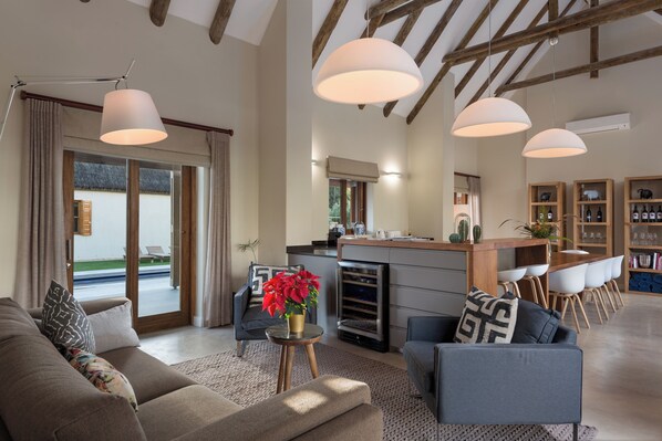 A spacious living area with a sofa, armchairs, and a coffee table. There is a kitchen area with a dining table and chairs, and a bar area with shelves of bottles.