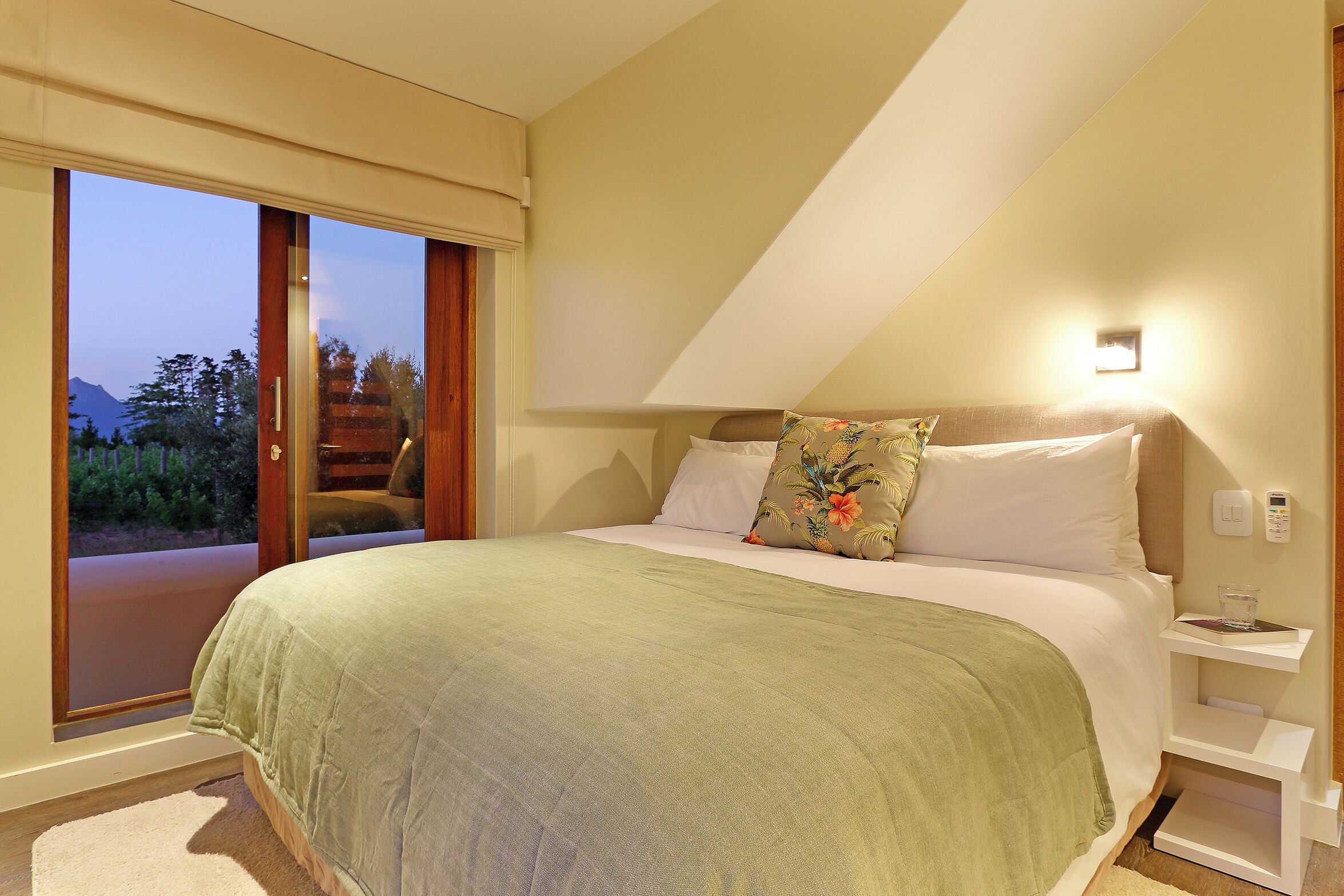 A bedroom with a large bed, a nightstand, a bedside table, a window with a view of the outdoors, and a door leading to a balcony.