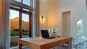 A wooden table with a laptop, a vase with flowers, and a chair in a room with large windows and a view of mountains.