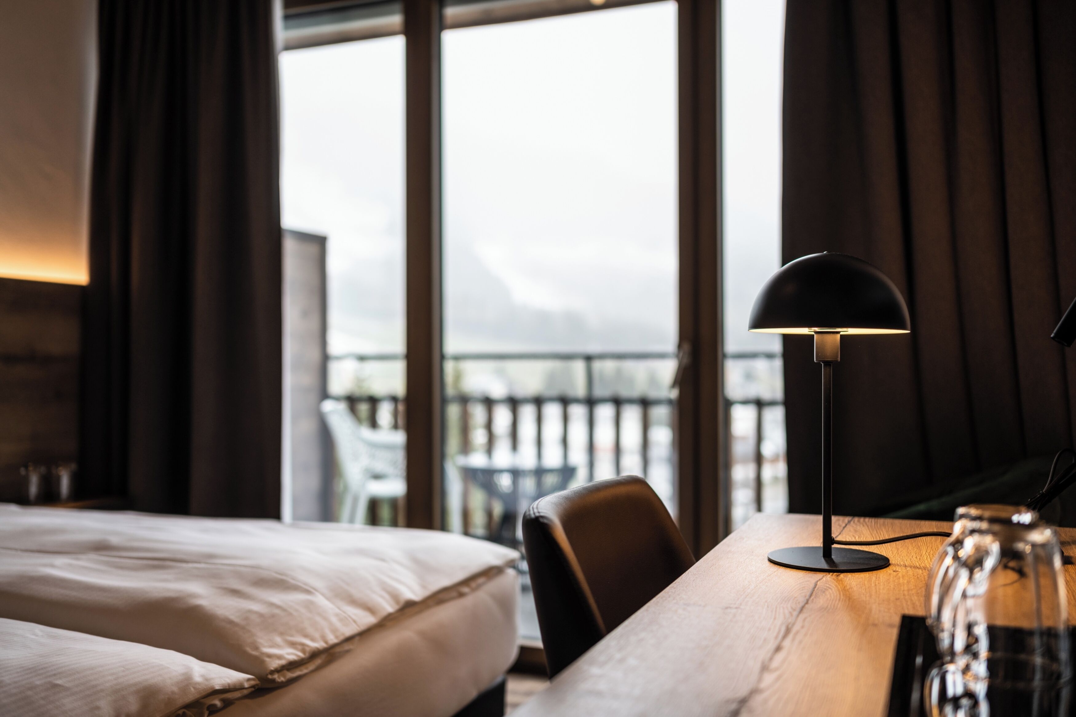 A hotel room with a bed, a desk with a lamp, and a view of the outdoors through large windows.