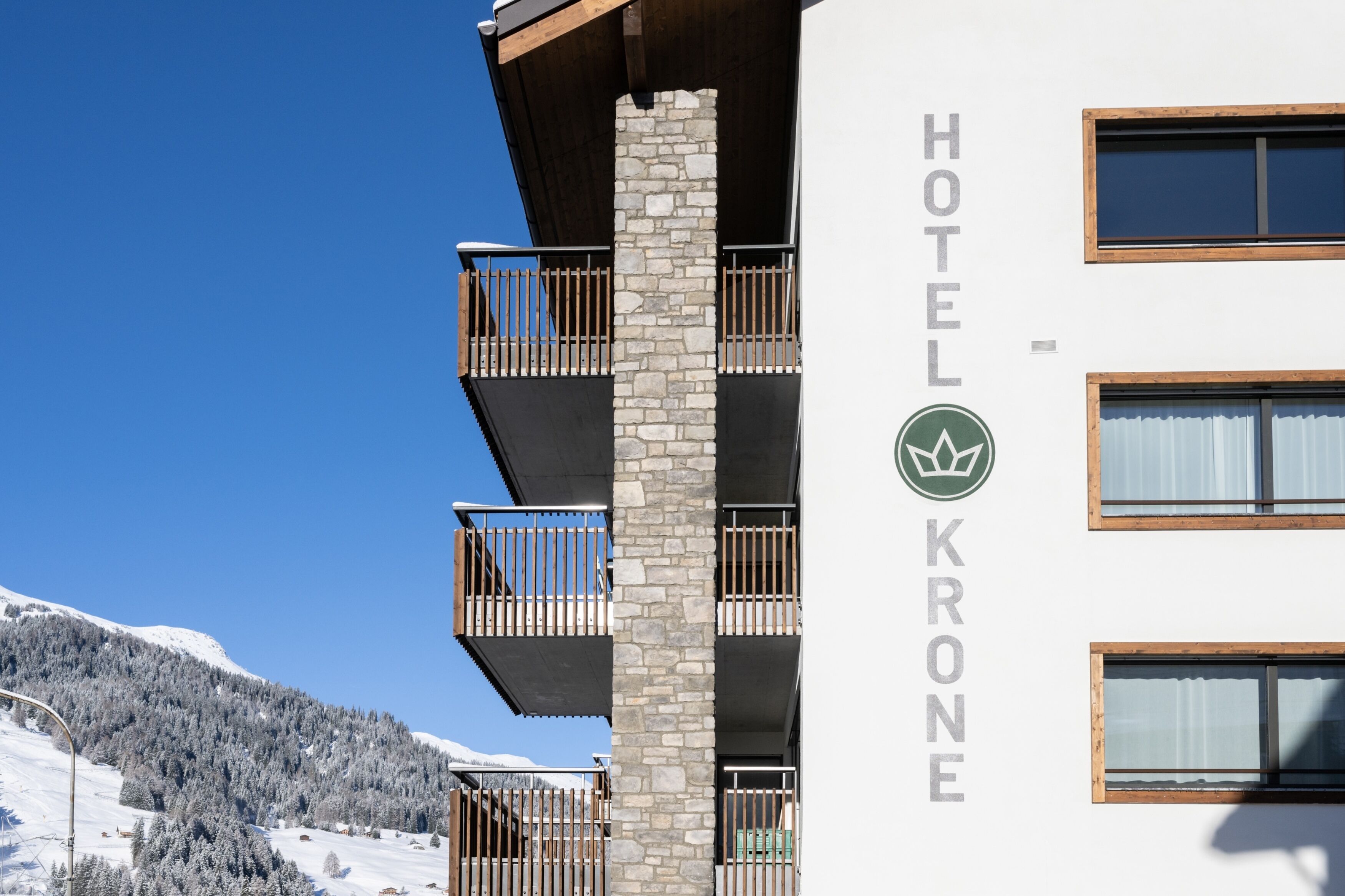 A hotel named "Hotel Krone" with a stone facade, multiple balconies, and a view of snow-covered mountains.