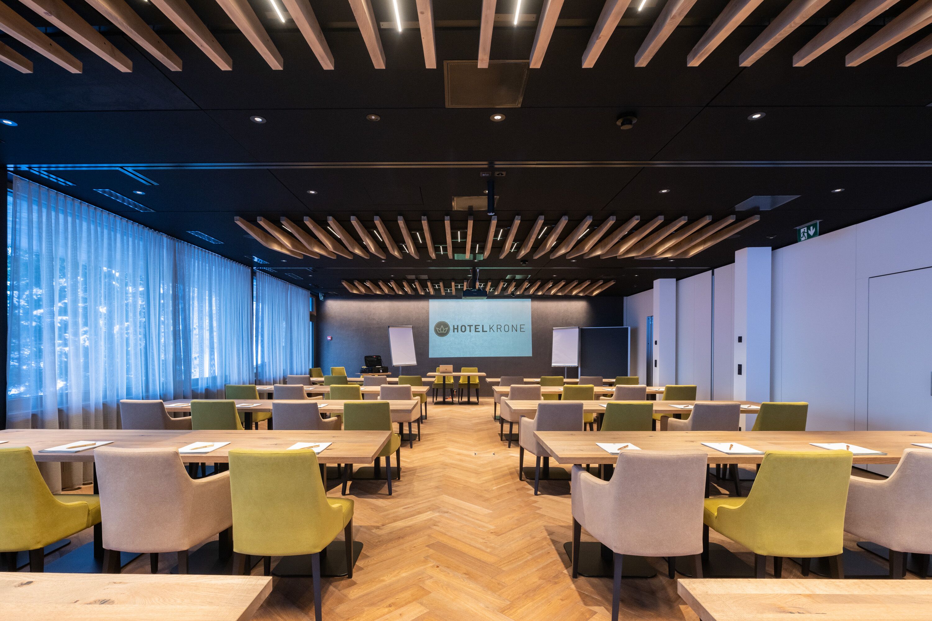 Una sala de conferencias con diseño moderno, que presenta una pantalla grande mostrando 'HOTELKRONE', varias mesas y sillas, y vistas a la ciudad a través de amplias ventanas.