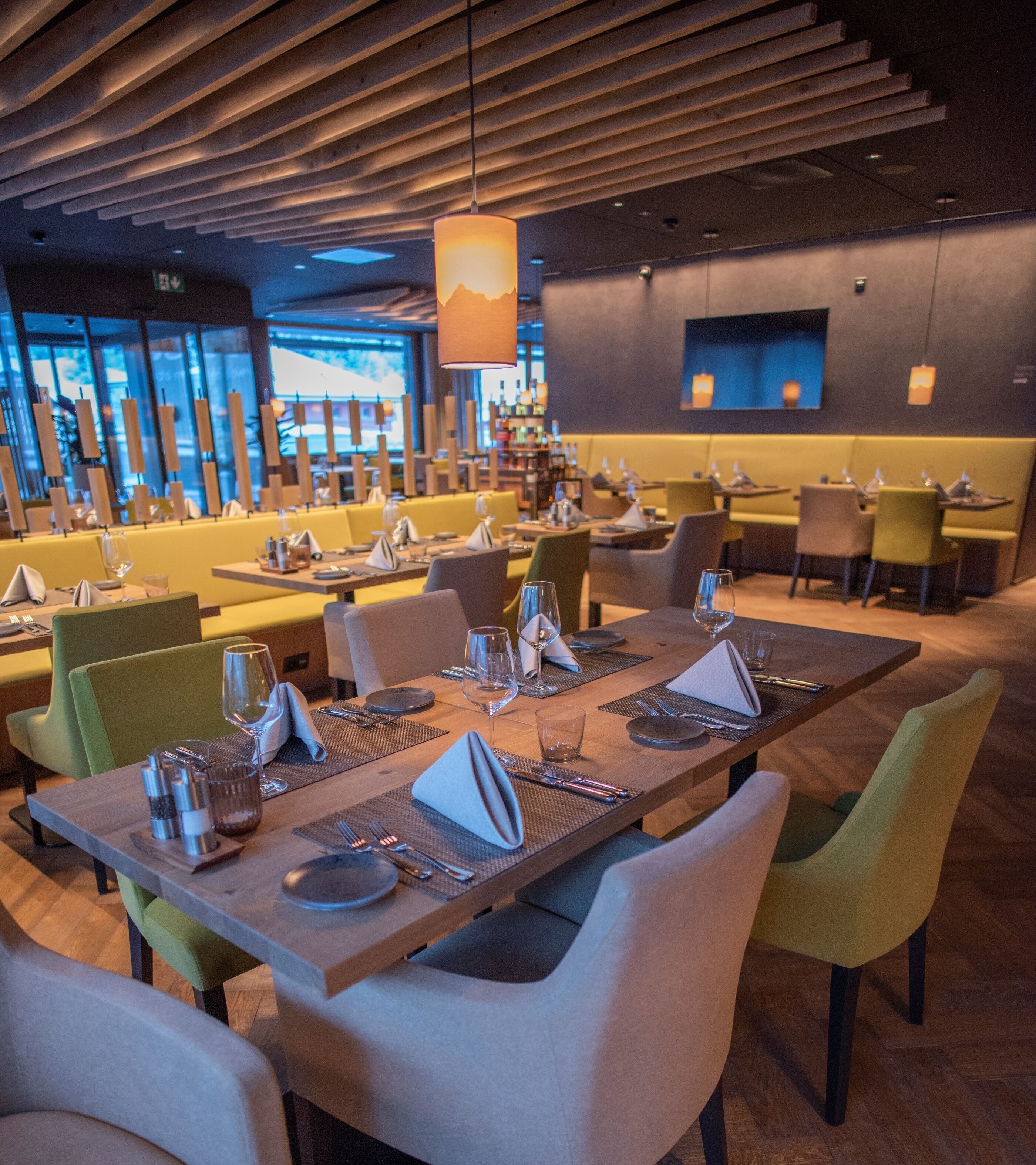 A modern restaurant with multiple dining tables and chairs, a wooden ceiling, and large windows in the background.