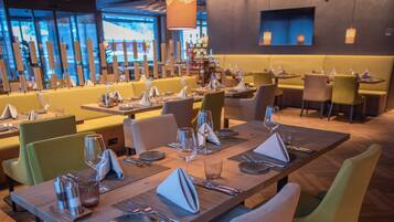 A modern restaurant with multiple dining tables and chairs, a wooden ceiling, and large windows in the background.