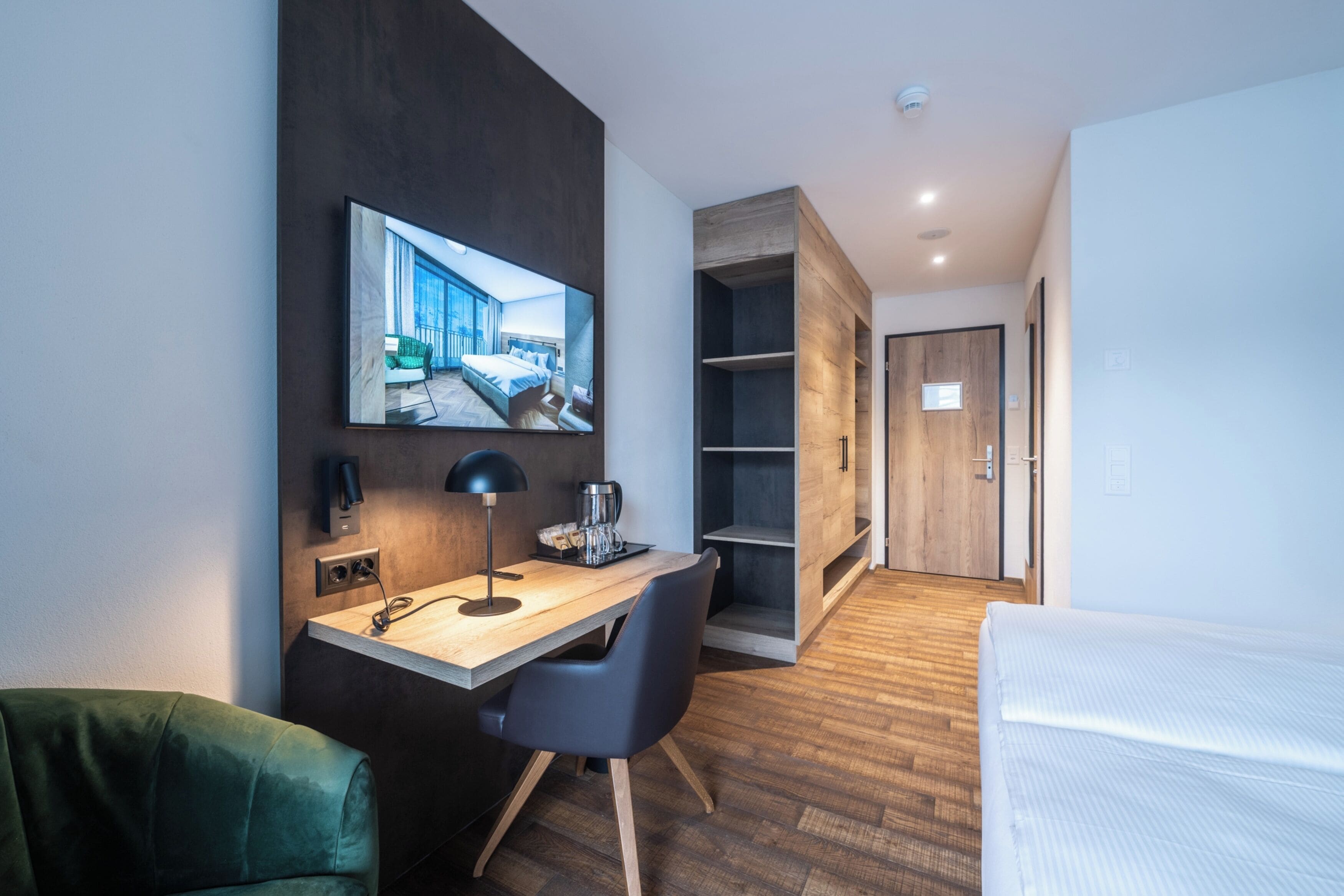 Una habitación de hotel moderna con un escritorio de madera, una cama, un televisor montado en la pared y un armario.
