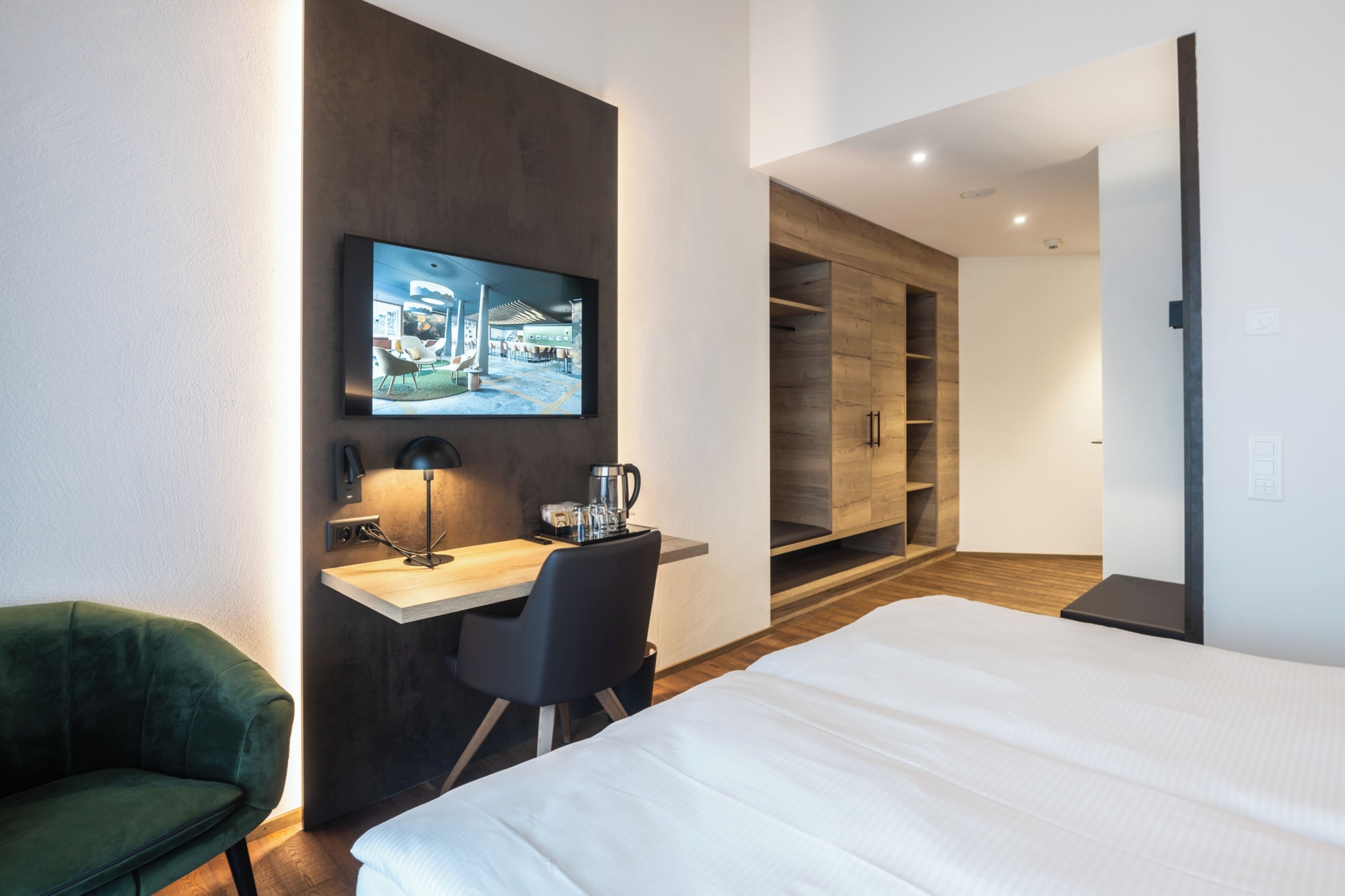 Una habitación de hotel moderna con una cama, un escritorio con silla, un televisor en la pared y un armario.