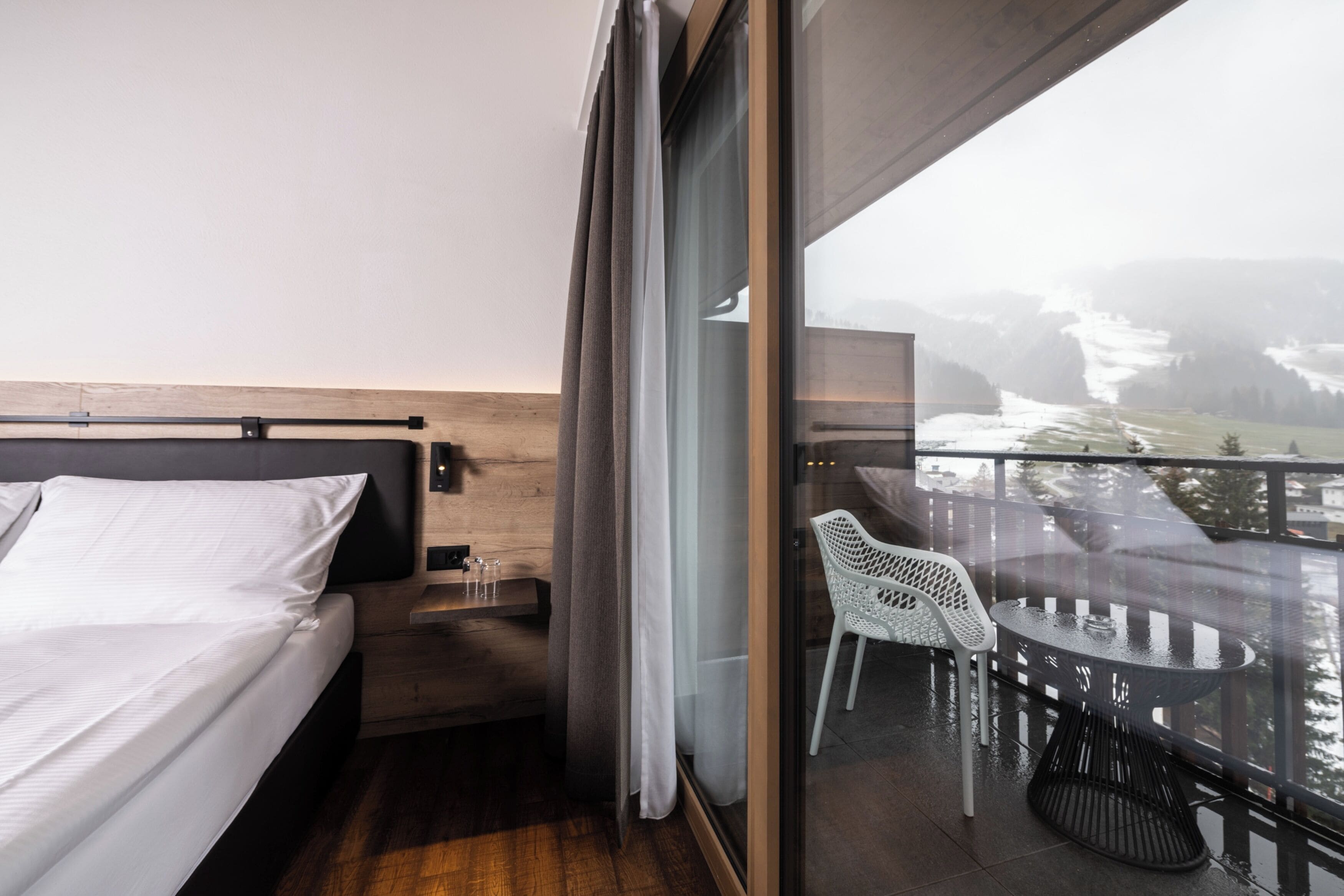 A modern hotel room with a bed, a small table, and a chair. The room has a balcony with a view of a snowy landscape.