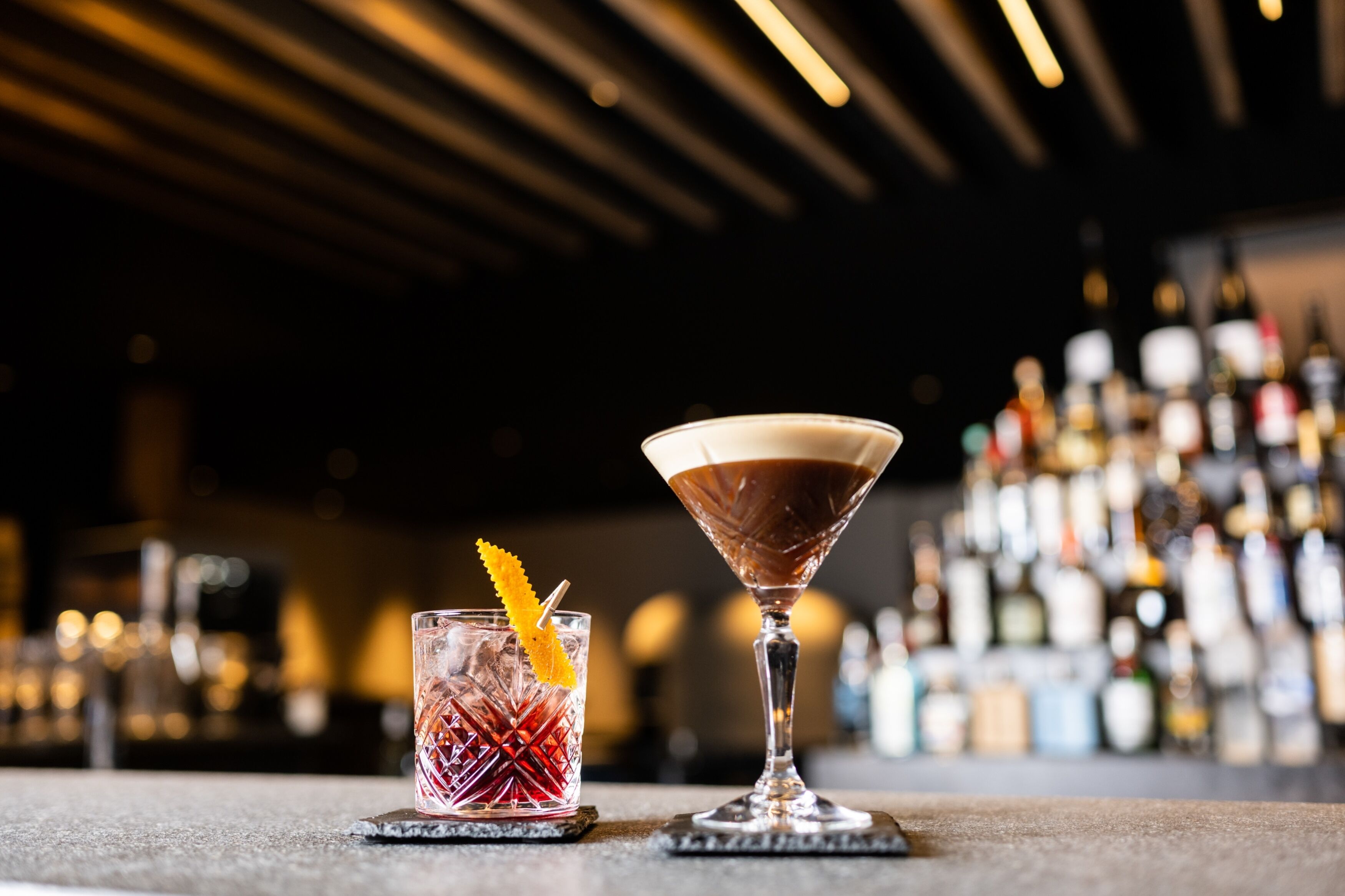 Twee cocktails op onderzetters aan een bar, met een wazige achtergrond van flessen.