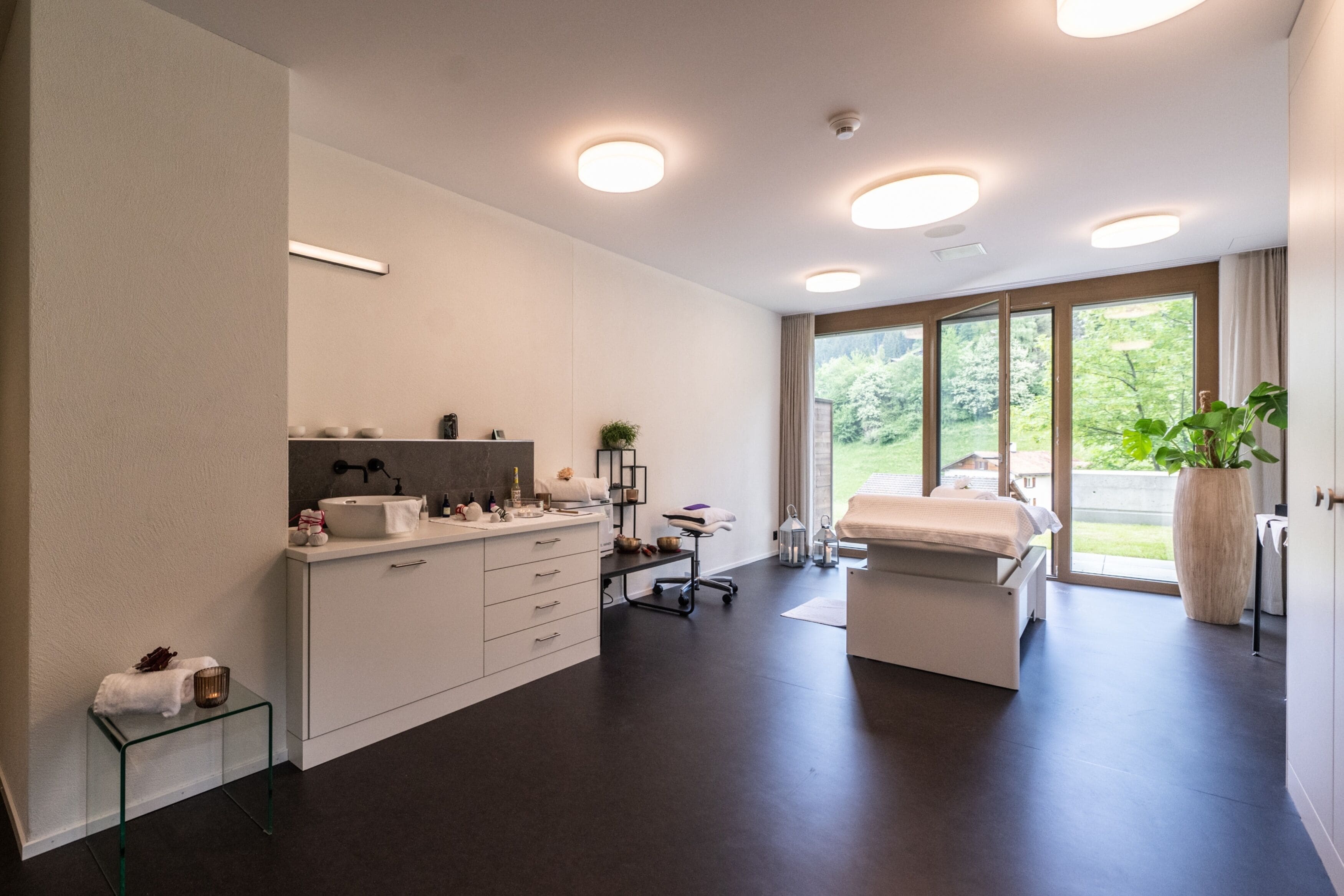 A modern bedroom with a large window, a bedside table, a bed, and a bathroom with a sink and mirror.