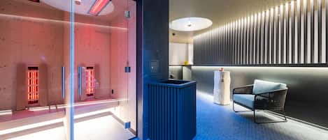 A modern spa area with a glass door entrance, a blue-tiled floor, a black trash bin, a white chair, and a wall with vertical slats.