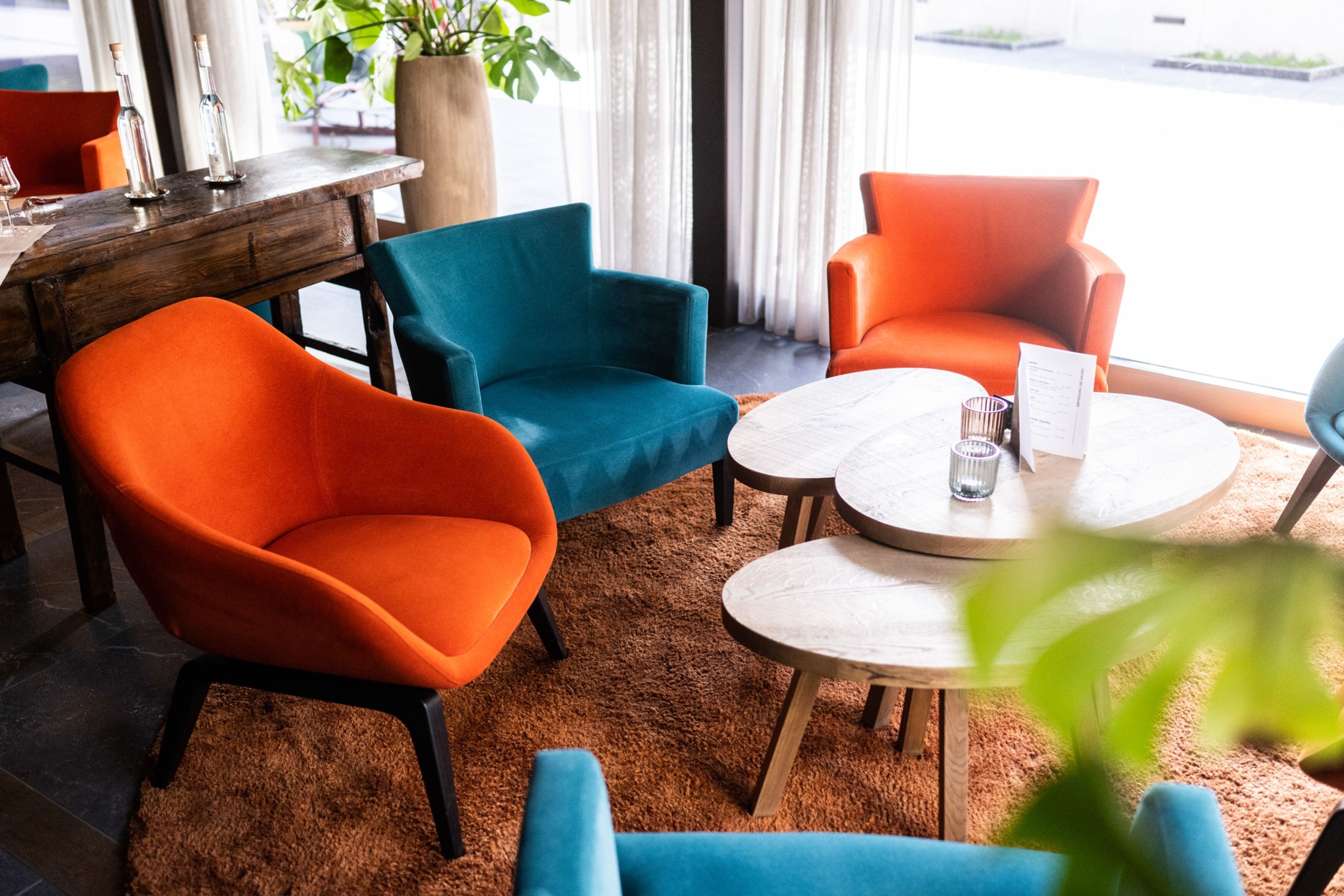 A modern lounge area with vibrant orange and teal chairs, a wooden table, and a large window with curtains.