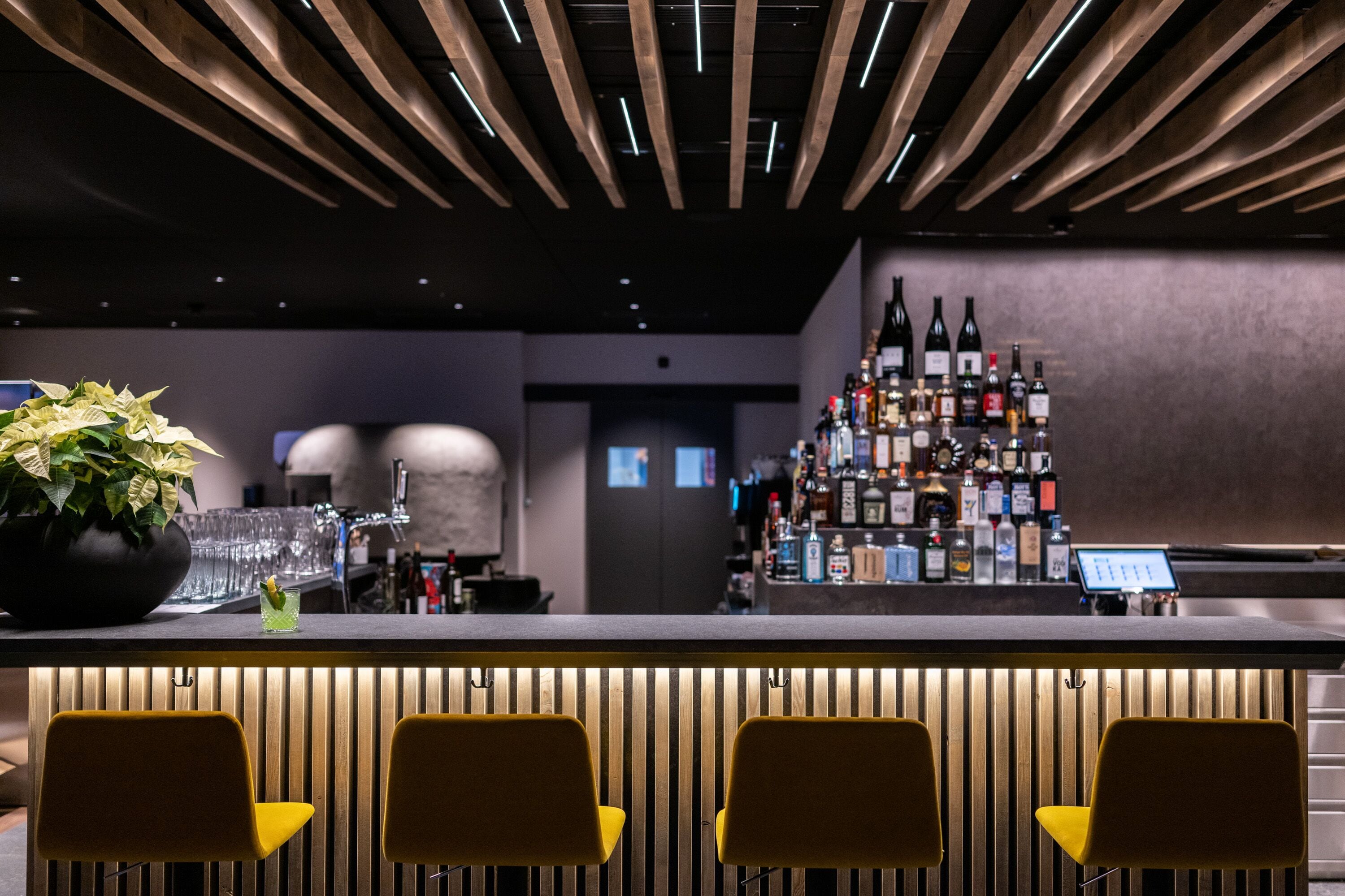A bar area with a wooden ceiling, a bar counter with multiple bottles, and yellow chairs.