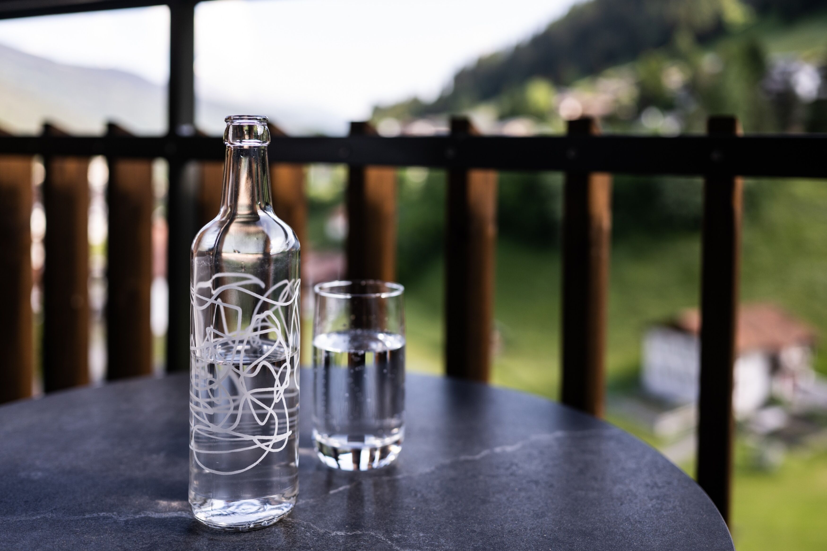 A bottle and a glass on a table with a view of greenery and hills in the background.