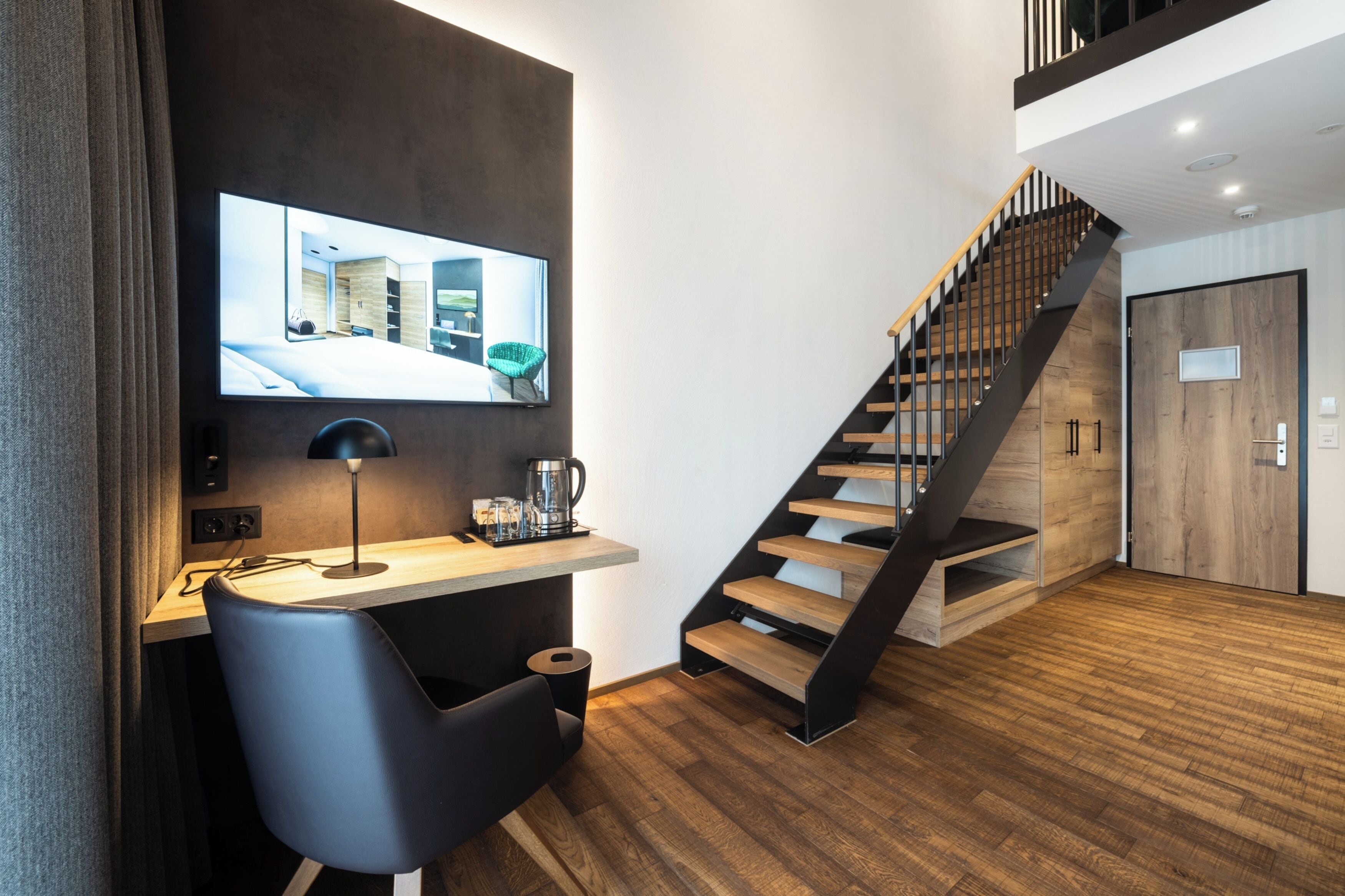Un intérieur moderne avec un escalier en bois, un bureau avec une chaise et un téléviseur fixé au mur qui diffuse une image d’une pièce.