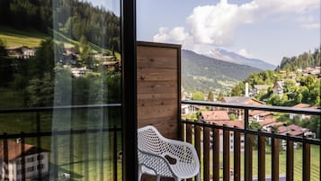 Un balcón con barandilla de madera, una silla de plástico blanca y vistas a un pueblo con casas y montañas al fondo.