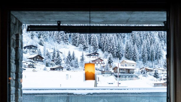 Une vue depuis une fenêtre d’un immeuble, donnant sur un paysage enneigé, avec des maisons et des arbres.