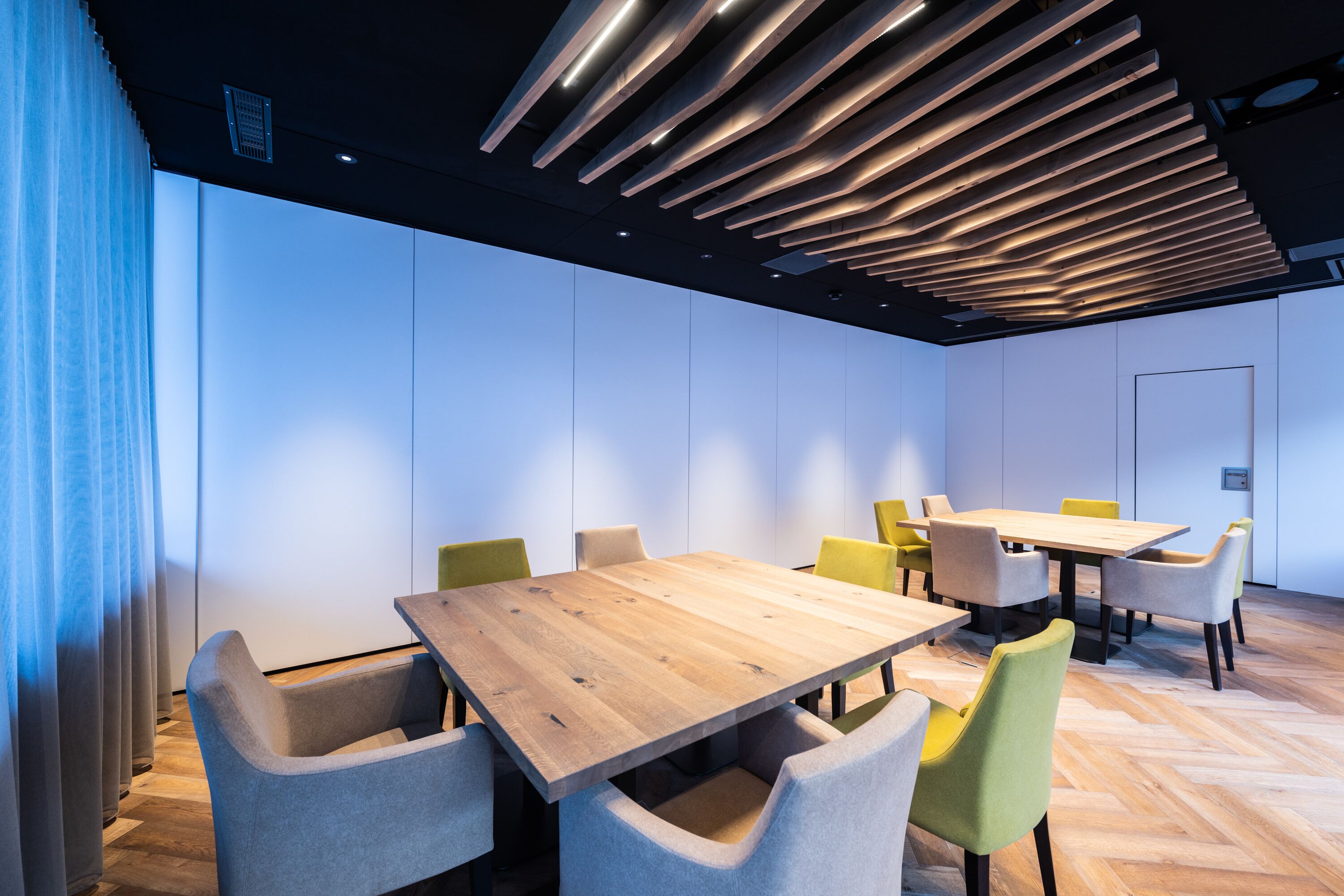 Una sala de conferencias moderna con mesas y sillas de madera, un diseño de techo con listones y un ventanal con cortinas.