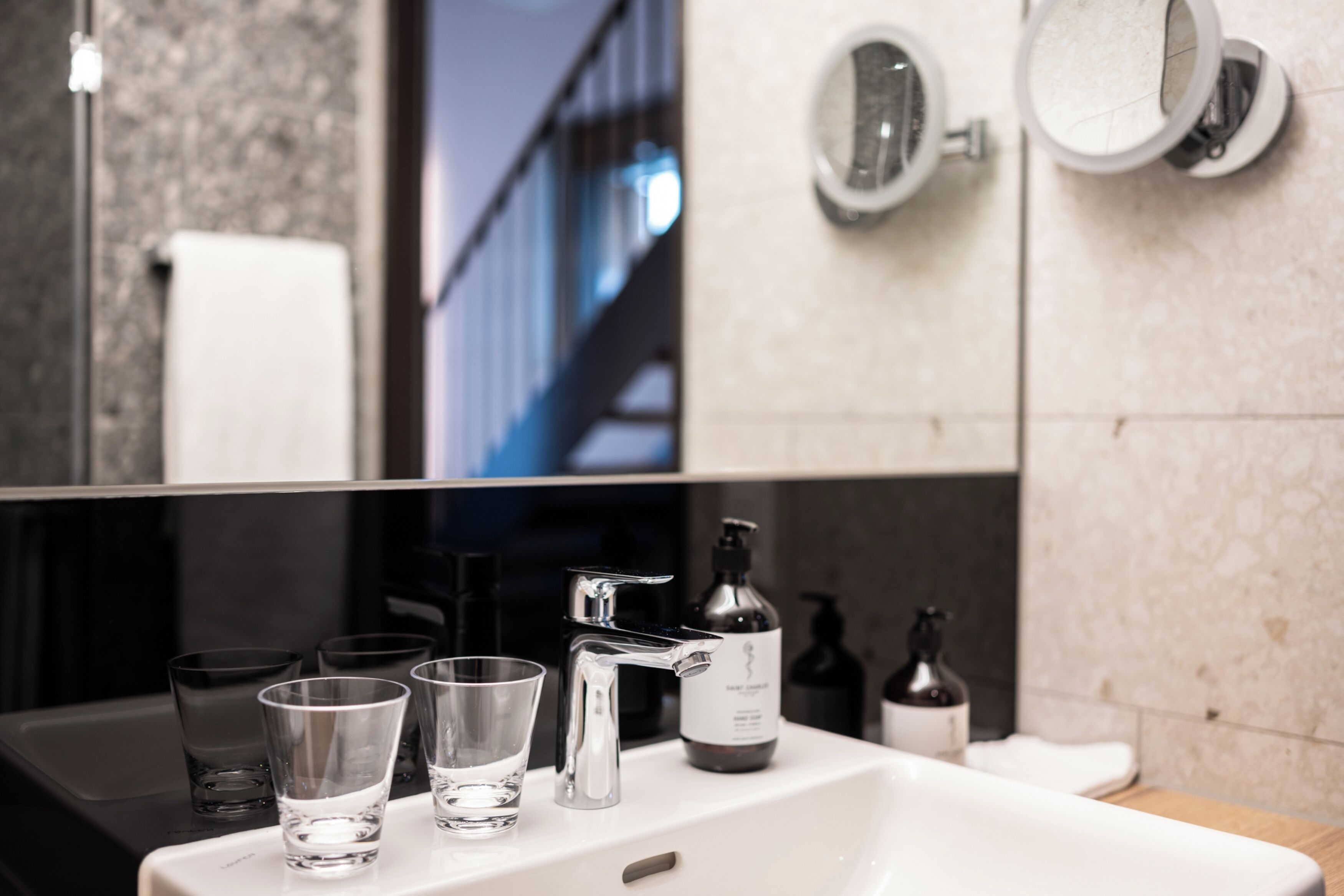 A bathroom with a white sink, two mirrors, and a large mirror above the sink. There are four clear glasses on the sink, a bottle of hand soap, and a bottle of lotion.