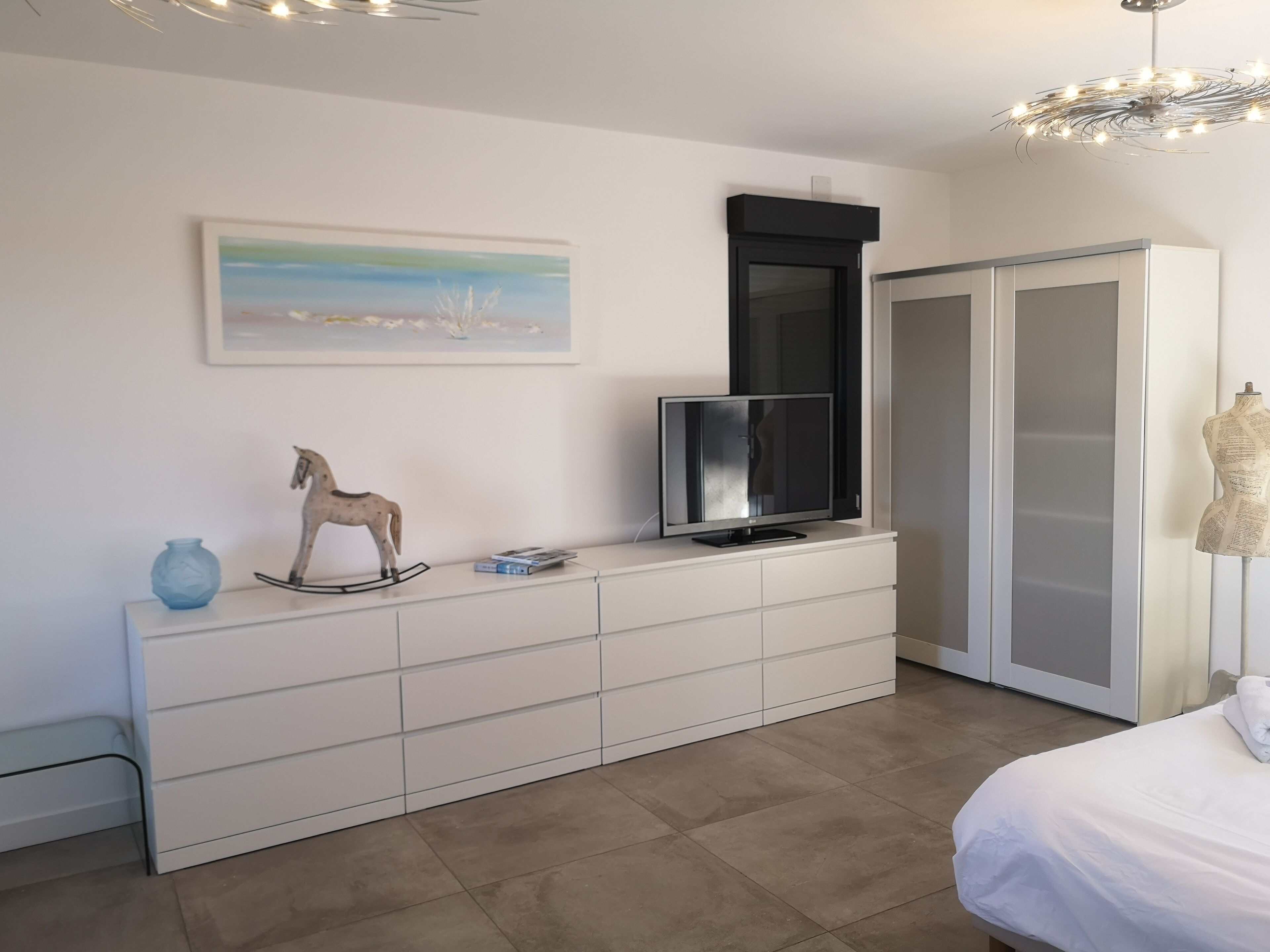 Ein modernes Schlafzimmer mit einer weißen Kommode, einem Fernseher, einer Pferdeskulptur, einem Gemälde und einem Kleiderschrank.